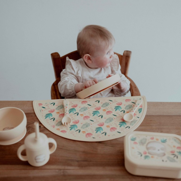 Silicone baby feeding plate beside stainless steel feeding ware and simple washing items on a neutral tabletop.