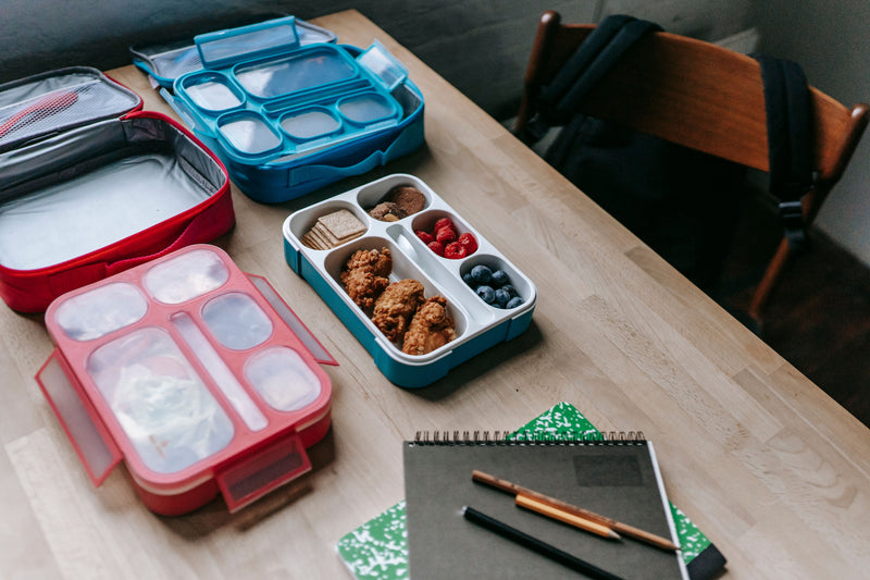 Two colourful school lunch boxes with berries and prepared food beside notebooks on a classroom table.