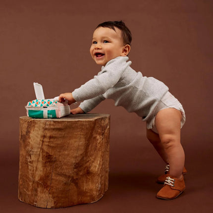 Baby standing on wooden stool next to Joonya Natural Baby Wipes 3-pack, showcasing product for sensitive skin with soothing ingredients.