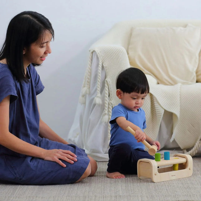Toddler plays with Plan Toys Pounding Bench, using a wooden mallet to hammer pegs, while mother watches, enhancing motor skills and engagement.