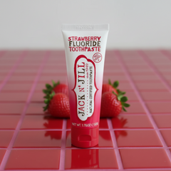 A tube of Jack N' Jill Strawberry Fluoride Toothpaste stands upright on a glossy red tiled surface with strawberries blurred in the background.
